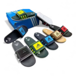 Multiple Adidas slide sandals in black, blue, green, tan and gold arranged around a blue Adidas box; product display image.