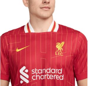Close-up of a red Liverpool FC football jersey with a yellow Nike swoosh, club crest, and Standard Chartered sponsor, featuring a white collar edge.