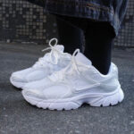 White chunky athletic sneakers tied with laces, worn with black socks and denim jeans on a pavement background