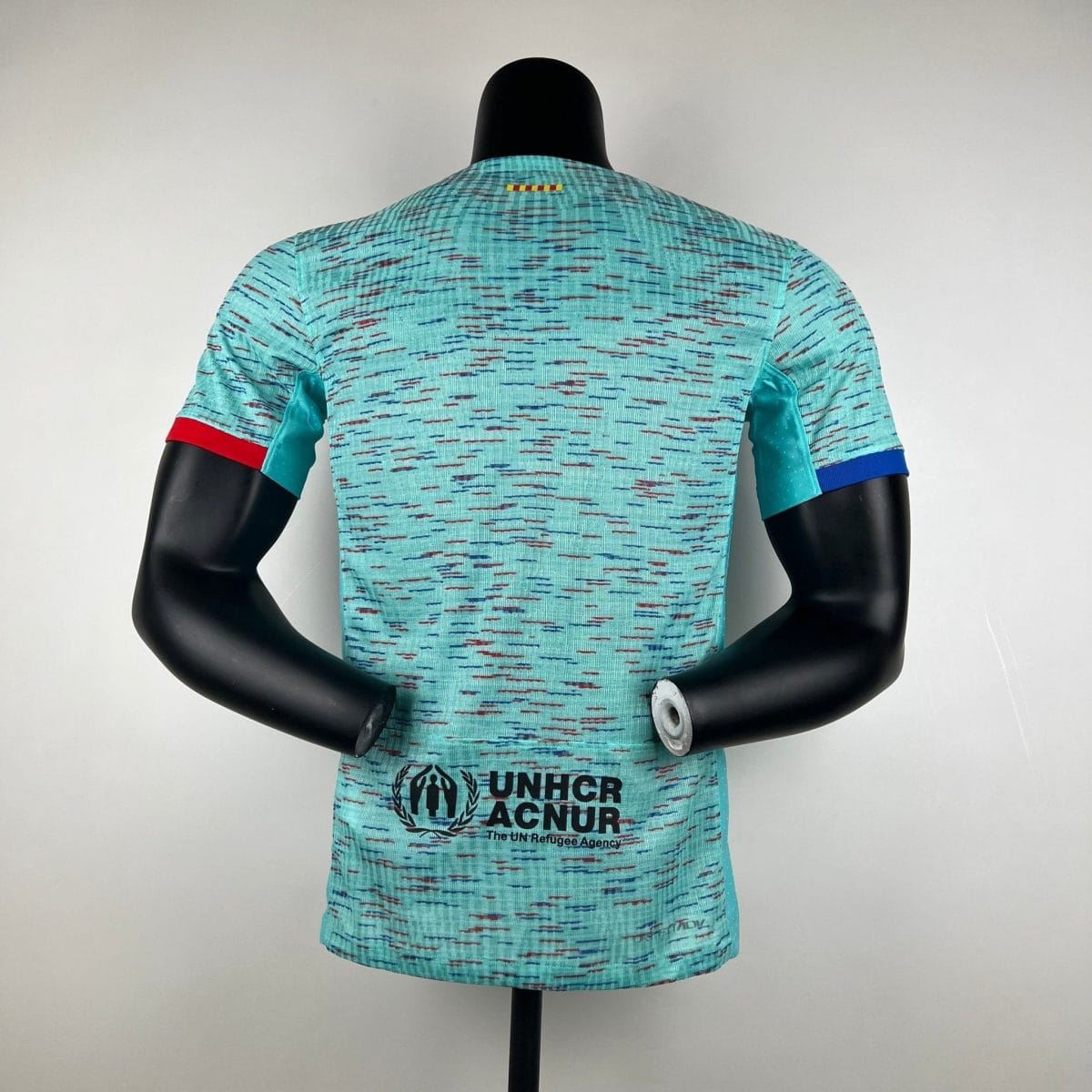 Back view of a turquoise patterned shirt on a mannequin with UNHCR ACNUR text visible at the hem.