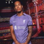 rn-image_picker_lib_temp_6f53b52f-b11c-47bb-964d-48d043efbcd1 | UAE SPORT STORE Man wearing a purple Liverpool FC jersey posing in a theater with red seats and decorative lighting in the background.