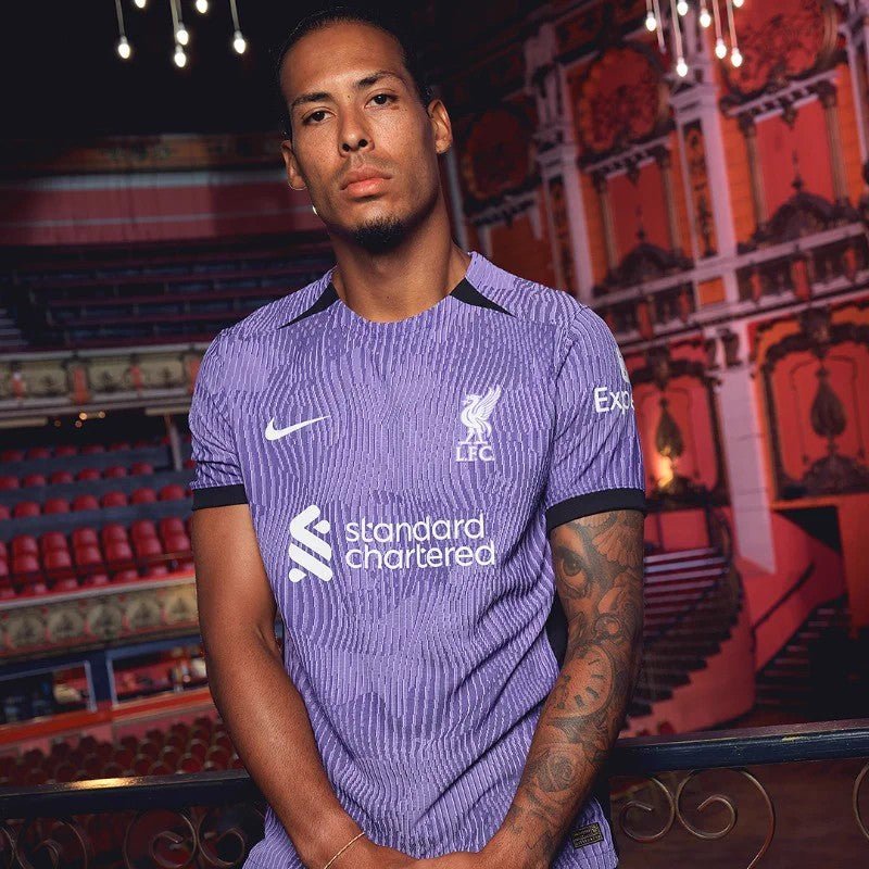 rn-image_picker_lib_temp_6f53b52f-b11c-47bb-964d-48d043efbcd1 | UAE SPORT STORE Man wearing a purple Liverpool FC jersey posing in a theater with red seats and decorative lighting in the background.