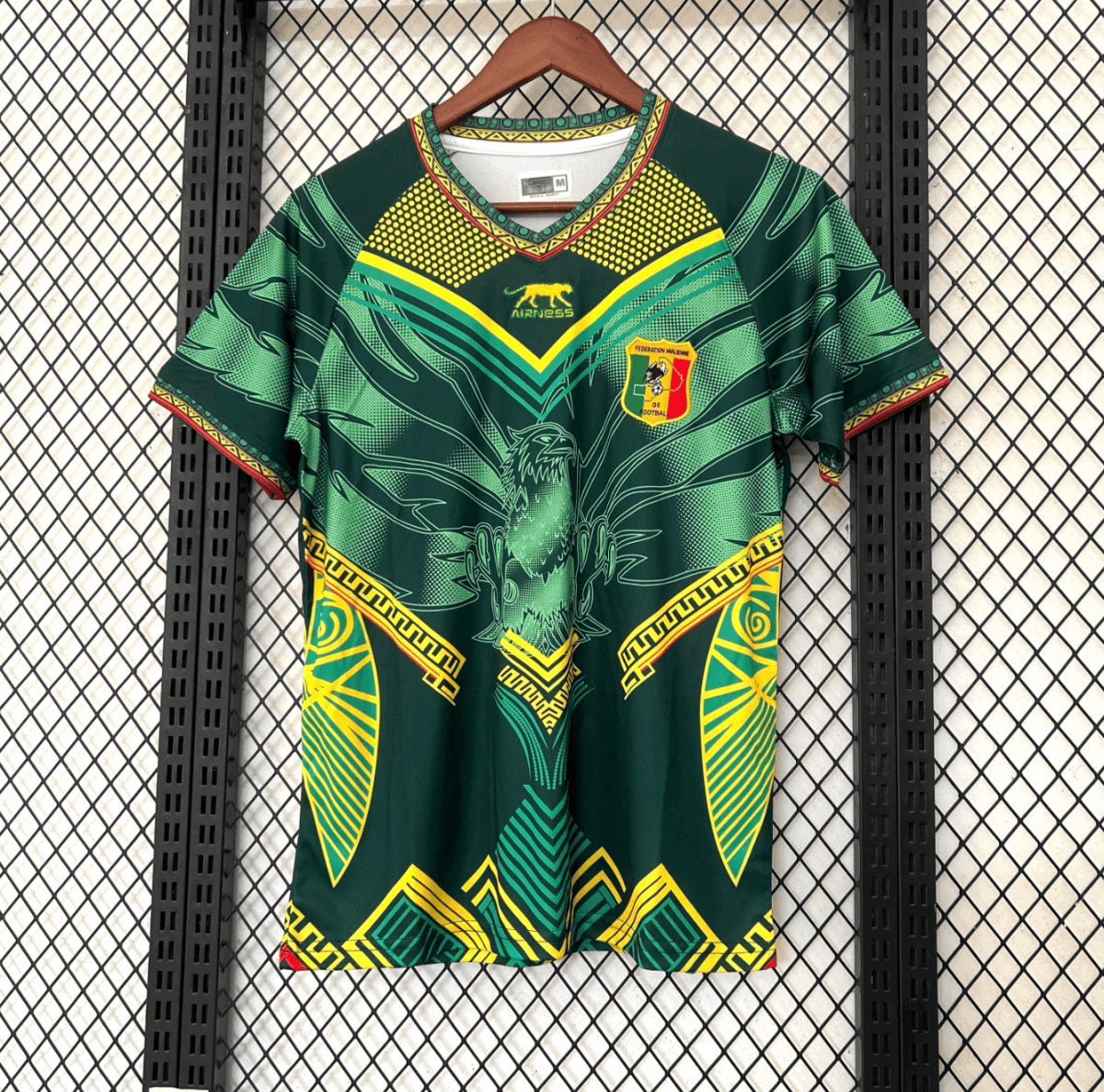 Green football jersey with yellow geometric patterns and an eagle emblem, Mali federation crest on the chest, hanging on a wooden hanger against a mesh background.