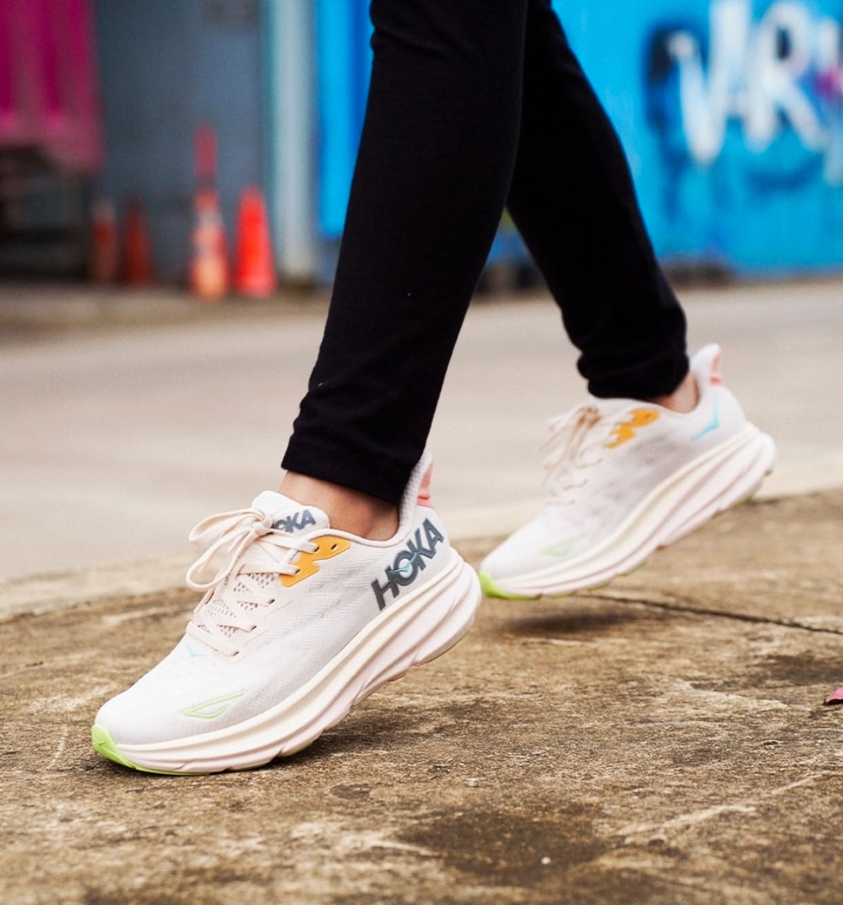 rn-image_picker_lib_temp_70d533a3-3ff8-4e54-8ff3-59ce39686263 Close-up of white HOKA running shoes stepping on a sidewalk, with black pants and a colorful graffiti backdrop.