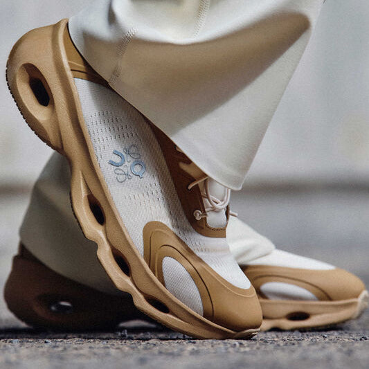 Close-up of a tan-and-white athletic sneaker stepping on pavement, highlighting the thick, sculpted sole with cutouts.