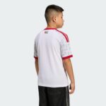 Back view of a person wearing a white sports shirt with red trim and a small flag detail on the upper back, against a light gray background.