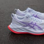 Close-up of lavender running shoes with purple accents resting on a dark asphalt surface, showing the side and sole details (FFTurbo+).