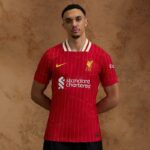 Portrait of a man wearing a red Liverpool FC jersey with Standard Chartered sponsor, standing with hands behind his back.