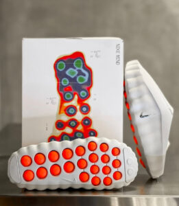 White Nike running shoe with bright orange circular outsole pods; box behind shows a multicolored foot-pressure heat map.