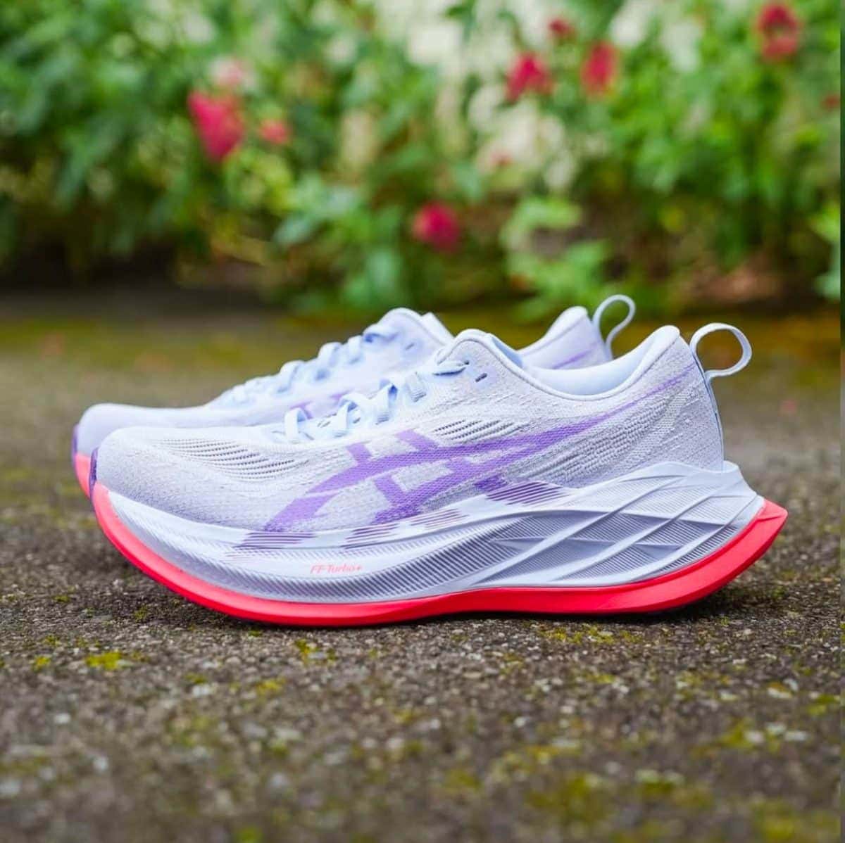 White running shoes with lavender accents and bright pink outsoles resting on a damp pathway with greenery in the background.