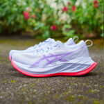 White running shoes with lavender accents and bright pink outsoles resting on a damp pathway with greenery in the background.