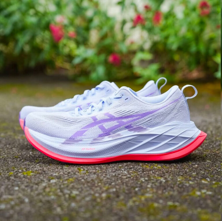 White running shoes with lavender accents and bright pink outsoles resting on a damp pathway with greenery in the background.