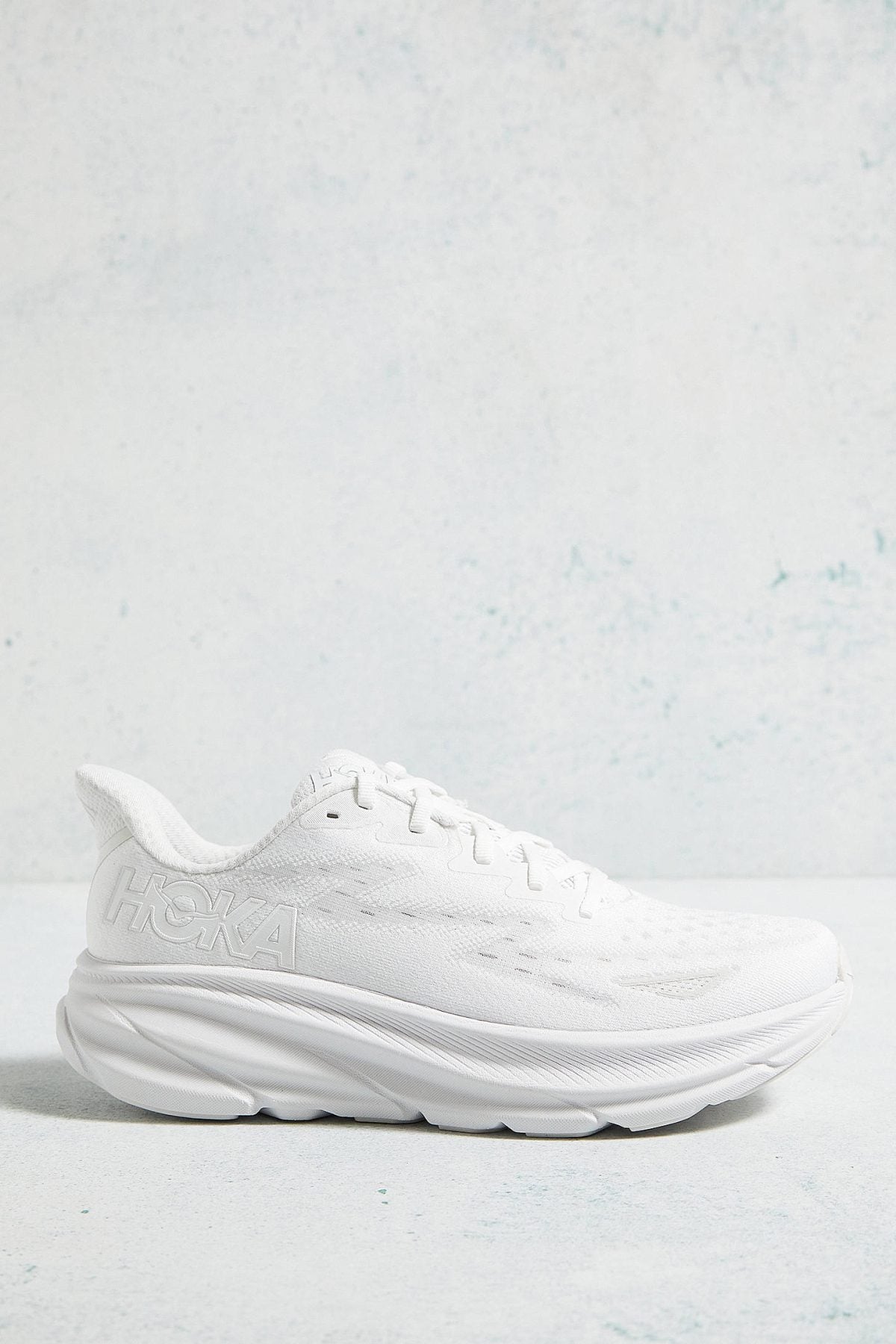 White Hoka running shoe with a chunky sole, set against a light blue textured background.