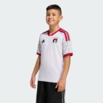 Young boy wearing a white UAE FA football jersey with red trim, standing against a light gray background.