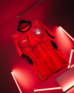 Red geometric-patterned football jersey with black trim, Puma logo, and club crest, hanging on a hanger under red neon lights on a dark background.