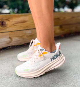 Close-up of pastel pink Hoka running shoes on a person’s feet, standing on a concrete surface with a wooden planter in the background, logo visible on the side.
