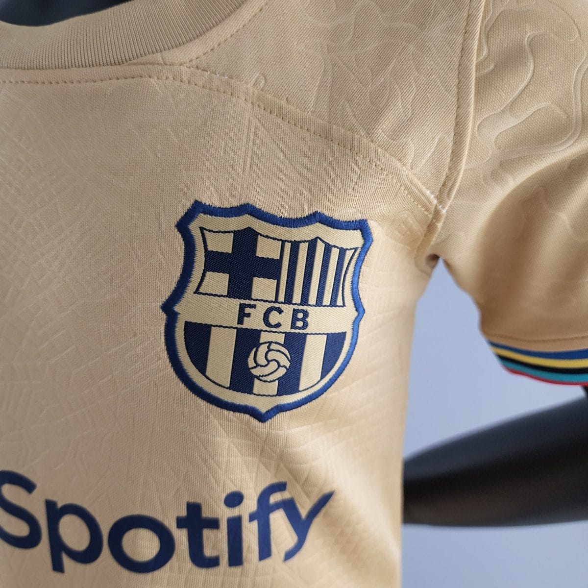 Close-up of a beige soccer jersey showing a blue-bordered shield crest with vertical stripes and the letters 'FCB' partially visible.