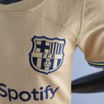 Close-up of a beige soccer jersey showing a blue-bordered shield crest with vertical stripes and the letters 'FCB' partially visible.