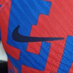 Close-up of red athletic jersey fabric with a blue stitched letter/shape, showing mesh texture and stitching detail