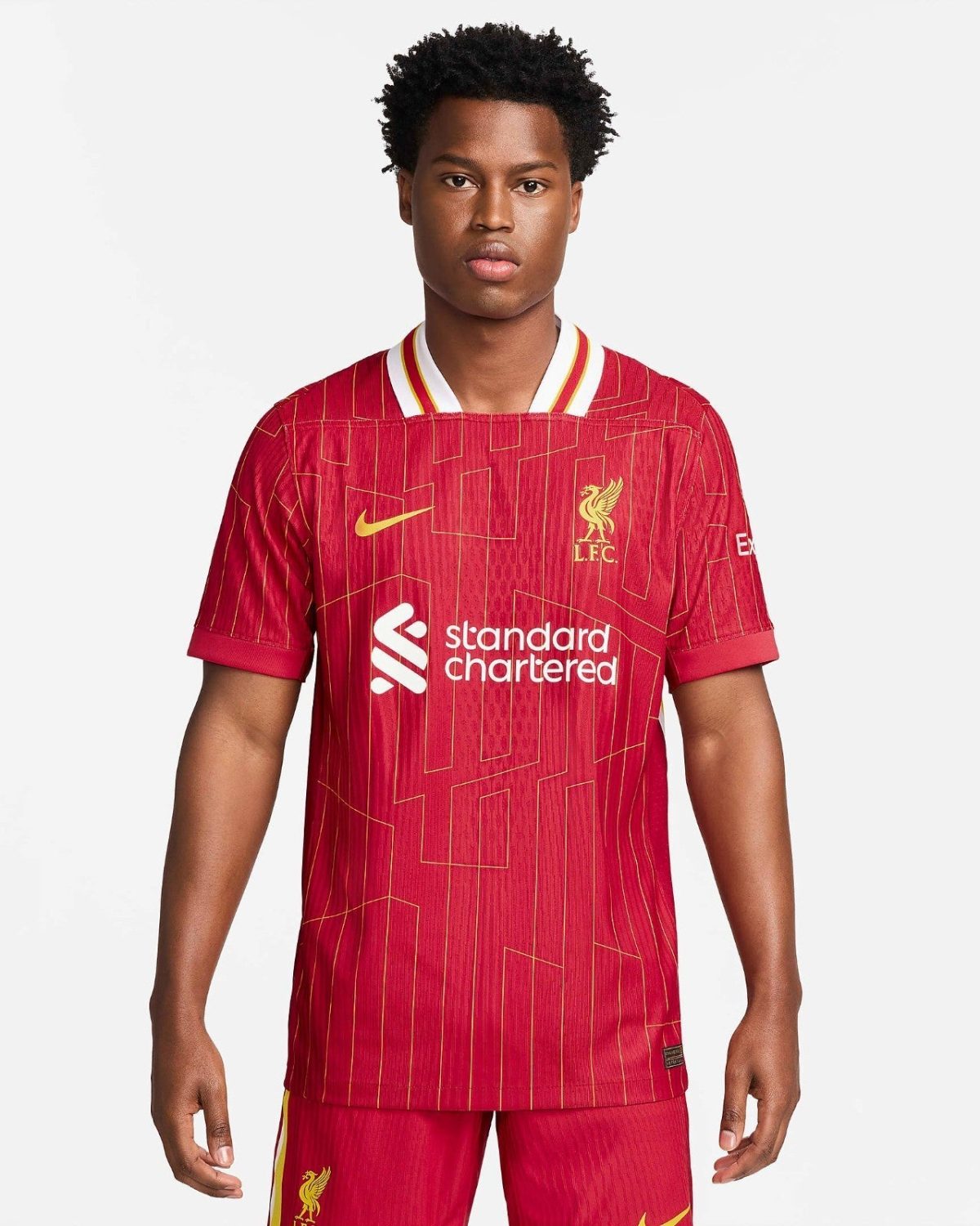Young man modeling a red Liverpool FC home jersey with yellow crest and sponsor logo, standing against a light background.