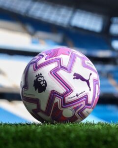 rn-image_picker_lib_temp_9863c8fa-4349-4be0-95d2-271c690acff2 | UAE SPORT STORE Soccer ball with pink-purple geometric patterns resting on grass in a stadium, featuring Premier League and Puma logos.