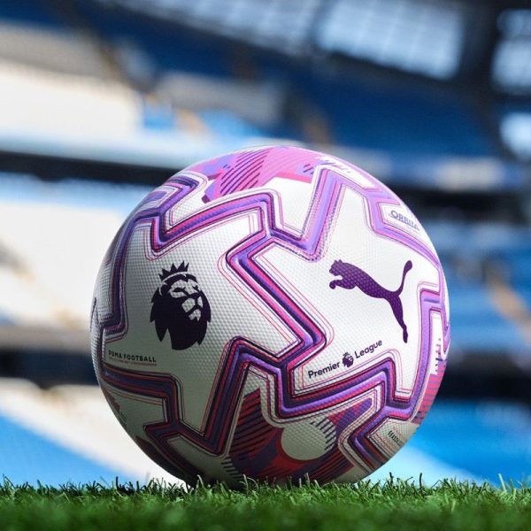 Soccer ball with pink-purple geometric patterns resting on grass in a stadium, featuring Premier League and Puma logos.