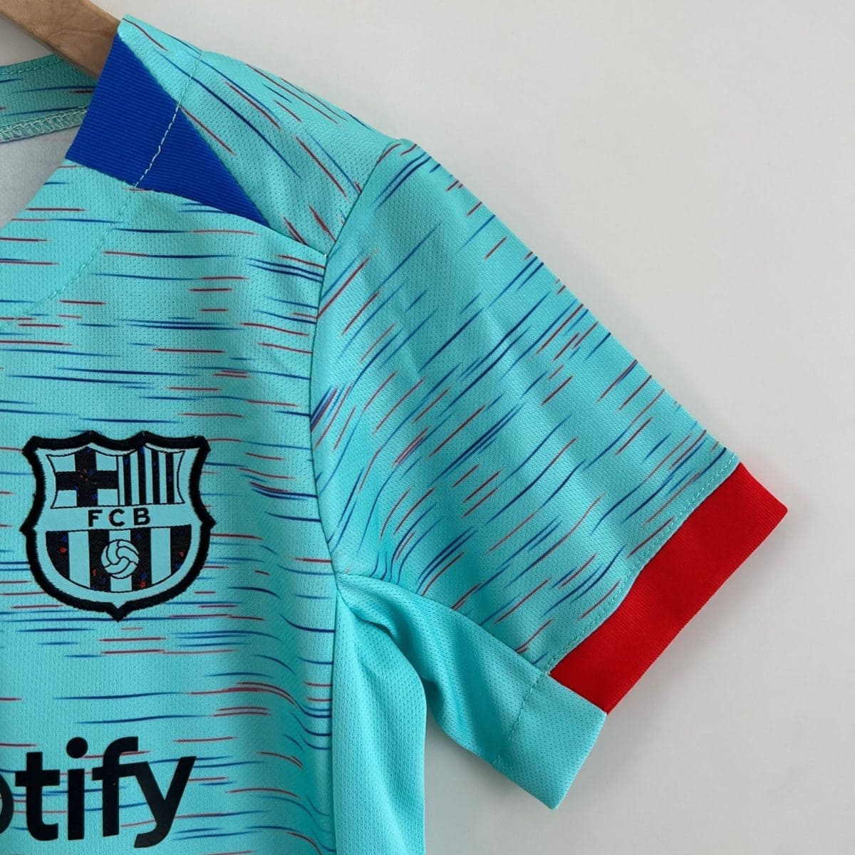 Close-up of a turquoise FC Barcelona jersey showing the club crest on the chest and a red sleeve edgeed cuff beneath, with diagonal line patterns.