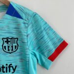Close-up of a turquoise FC Barcelona jersey showing the club crest on the chest and a red sleeve edgeed cuff beneath, with diagonal line patterns.