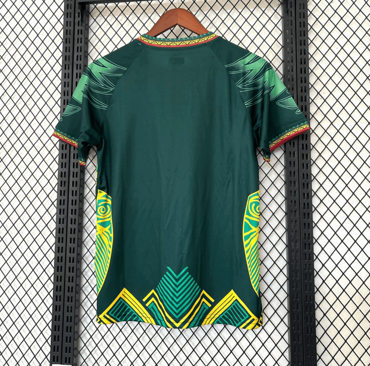 Back view of a dark green dress with bright yellow and turquoise geometric patterns along the hem and sides, hanging on a wooden hanger against a metal grid backdrop.