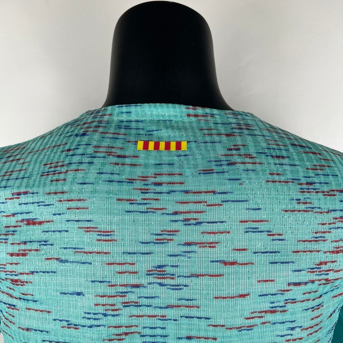 Back view of a black mannequin head and shoulders wearing a turquoise patterned fabric with a small yellow and red striped label on the chest.