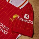 Red Liverpool FC jersey with yellow crest and Expedia sponsor logo on a wooden surface, showing team branding and design lines.
