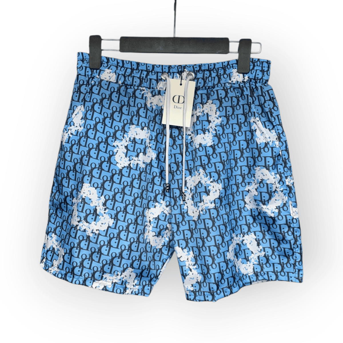 Blue shorts featuring a repeating Dior monogram pattern with white paint splatter details, displayed on a hanger with a tag attached