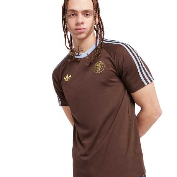 Young man with brown braided hair wearing a brown Adidas jersey with light blue stripes, standing with hands behind his back.