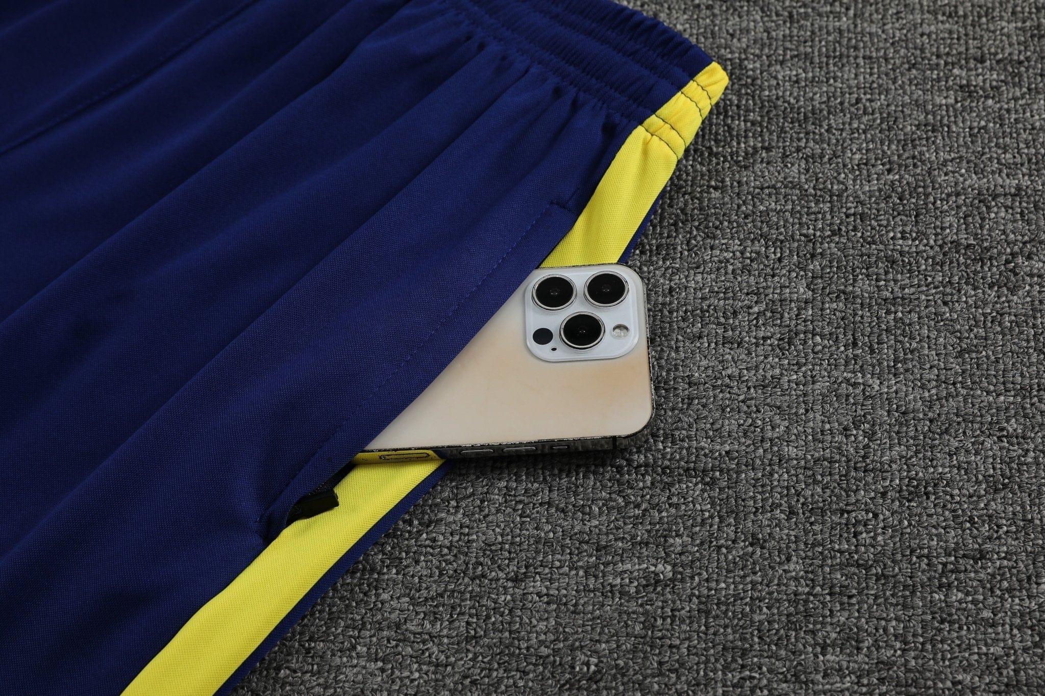 rn-image_picker_lib_temp_b8f34d8b-82f6-4b45-afb2-7185c03a542d | UAE SPORT STORE Close-up of a smartphone partially tucked under a blue and yellow gym jacket on a gray carpeted floor, shot from above.