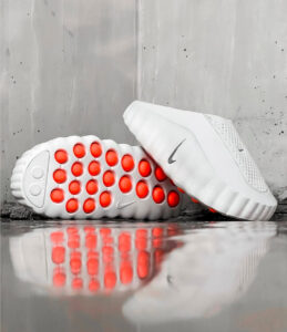 White athletic shoe with orange circular grip pods on the sole, shown on its side with a reflective surface nearby.