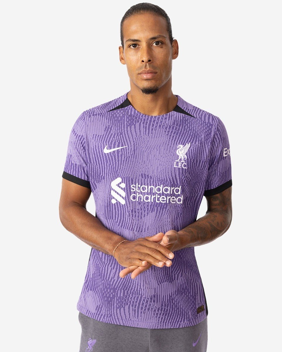 rn-image_picker_lib_temp_bffafafe-9652-4986-afda-9a626368f05c | UAE SPORT STORE Male model wearing a purple Liverpool FC jersey with Standard Chartered sponsor and club crest, standing with hands clasped.