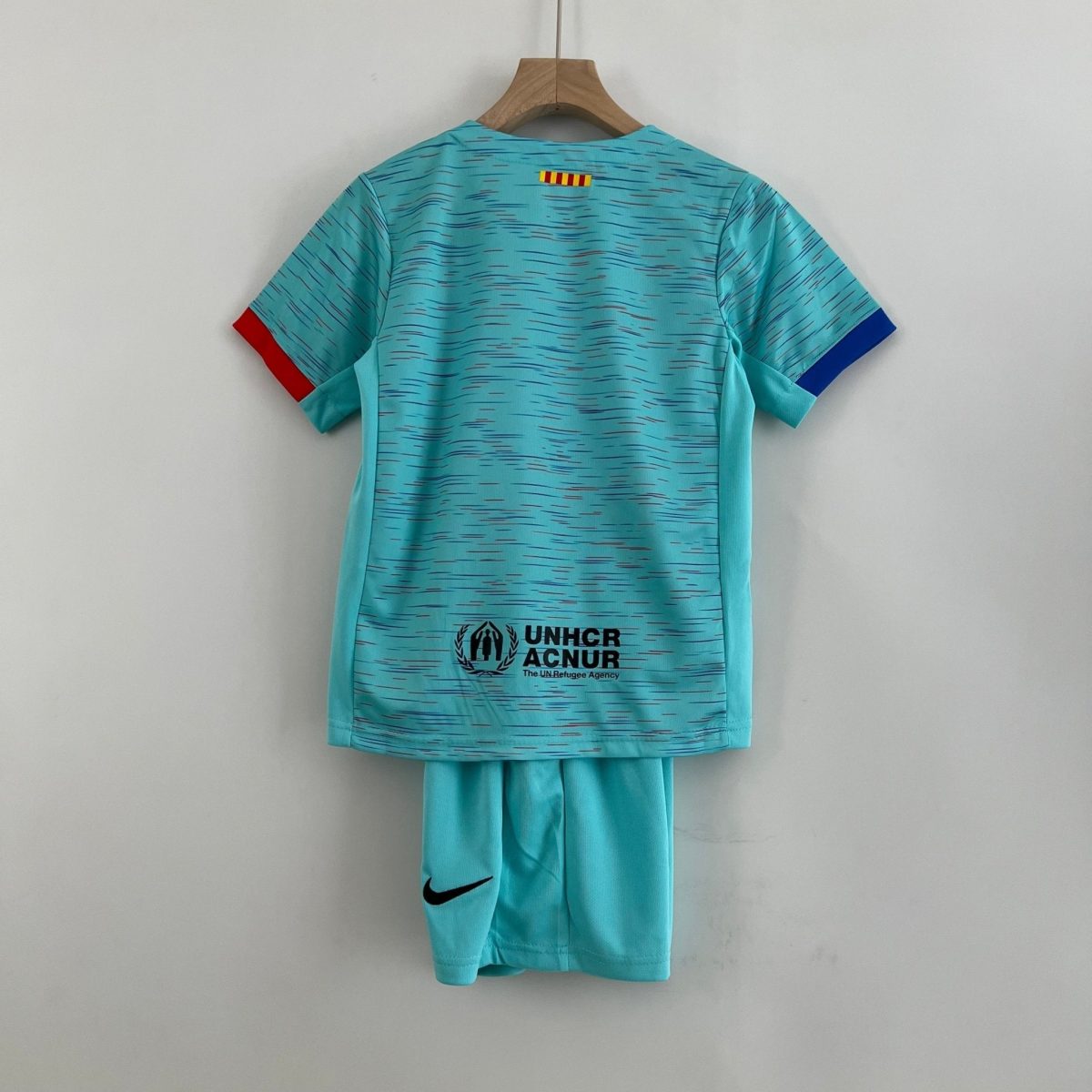 Turquoise sports jersey on a wooden hanger, back view with UNHCR/ACNUR logo and red/blue sleeve cuffs.