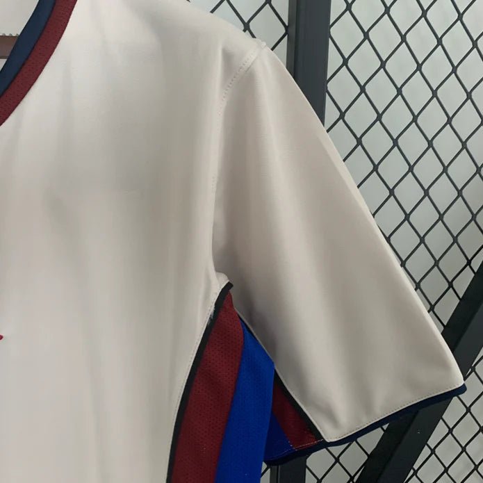 rn-image_picker_lib_temp_cad3615b-0ddf-4c17-9cf4-f1e6cfebdfc6 | UAE SPORT STORE Close-up of a white sports jersey with red and blue trim hanging beside a chain-link fence.