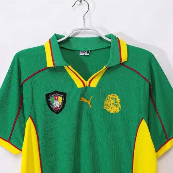 Green Cameroon national team football jersey on a hanger, with red and yellow trim and sponsor PUMA logo on the chest side. Approachable, informative.