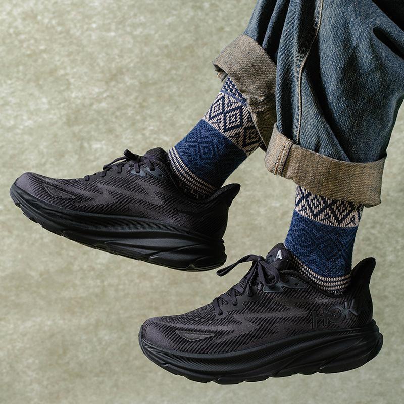 rn-image_picker_lib_temp_cc8ae230-c2b7-42f2-8d1c-9ce2b35d1e47 Black athletic sneakers with dark patterned socks and rolled denim cuffs against a concrete background.
