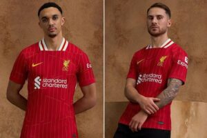 Two men wearing red Liverpool FC home jerseys stand against a brown textured wall; left man has hands behind his back, right man sits with arms crossed.