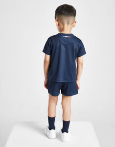 Back view of a young boy in navy athletic outfit (t-shirt and shorts) with white sneakers, standing on a white surface. Near the neck is a small flag detail on the shirt.