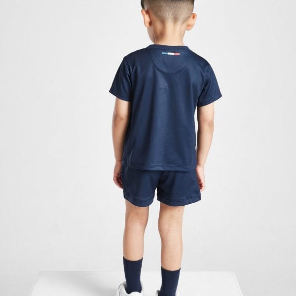 Back view of a young boy in navy athletic outfit (t-shirt and shorts) with white sneakers, standing on a white surface. Near the neck is a small flag detail on the shirt.