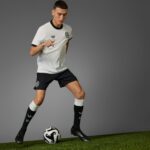 rn-image_picker_lib_temp_d6c32434-e0d4-4c77-acfe-ffde95ea96b0 | UAE SPORT STORE Soccer player in a white jersey and black shorts dribbles a soccer ball on a grassy field against a gray background.