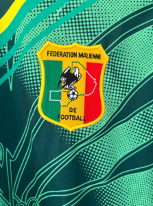 Embroidered shield logo of the Fédération Malienne de Football: green, yellow, red vertical stripes, black eagle with a football, on a teal fabric background.