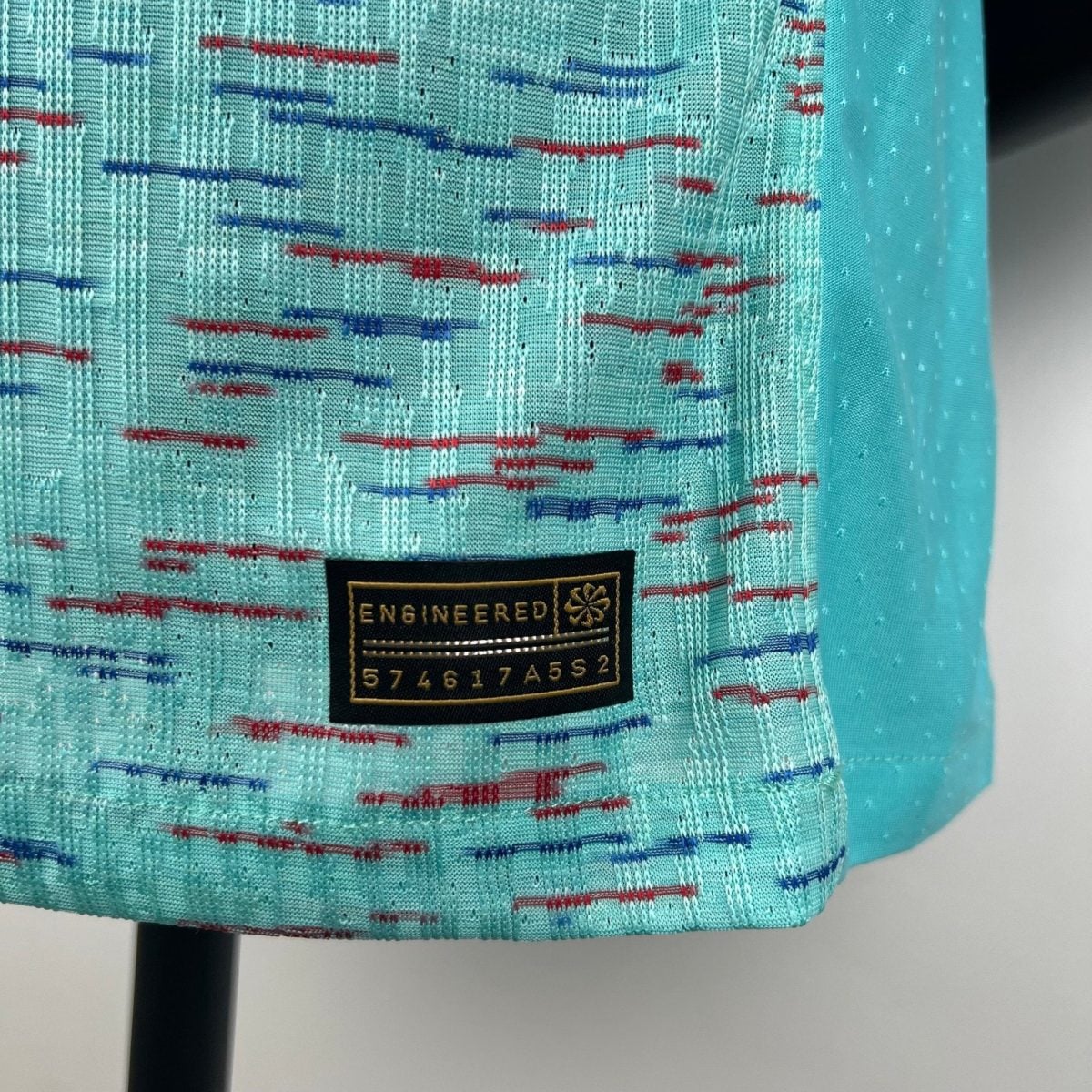 Close-up of turquoise woven fabric with red and blue stitched lines and a black label reading 'ENGINEERED'.