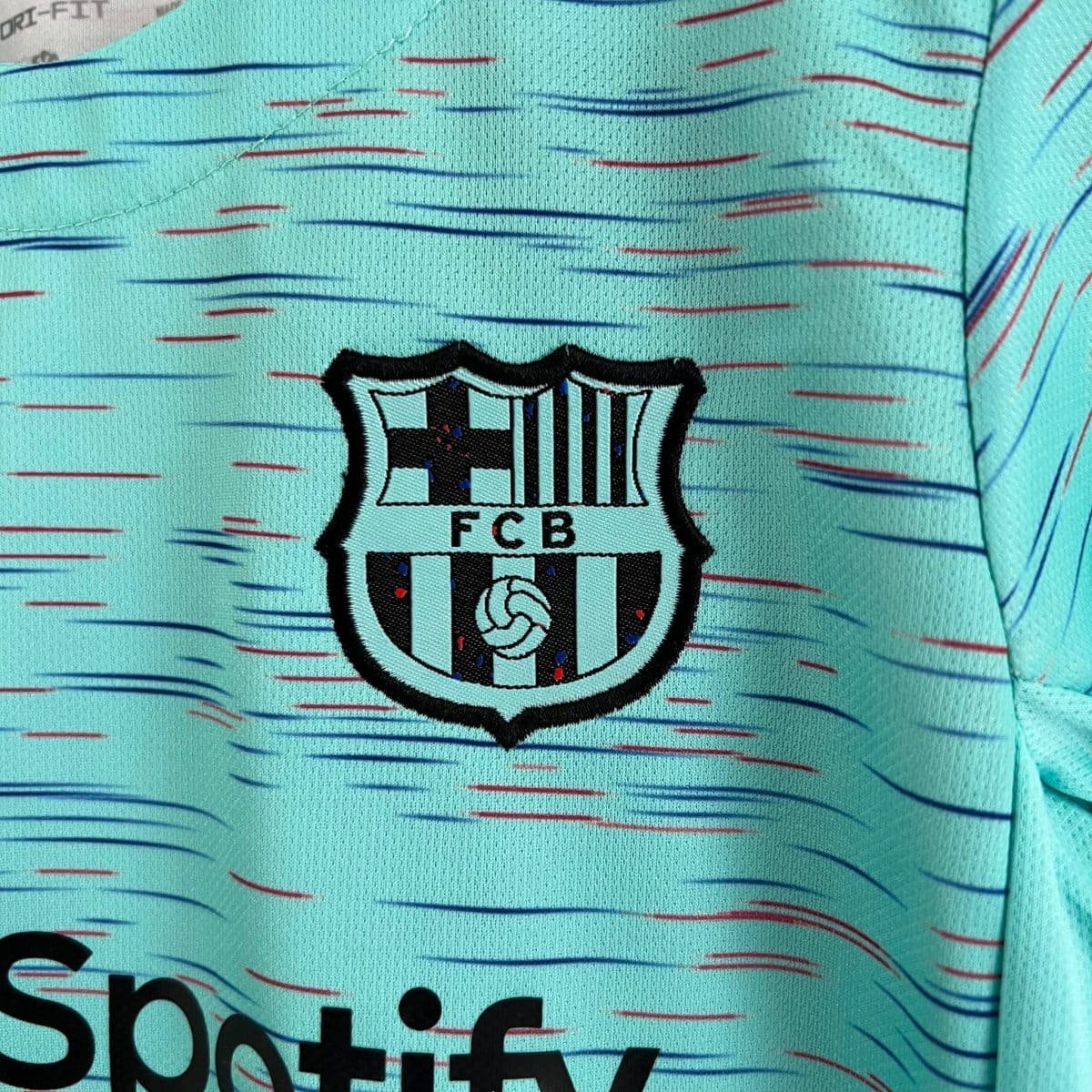 Close-up of teal fabric with a stitched shield logo reading 'FCB', resembling a soccer club crest with black and white stripes and a small soccer ball.