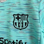 Close-up of teal fabric with a stitched shield logo reading 'FCB', resembling a soccer club crest with black and white stripes and a small soccer ball.
