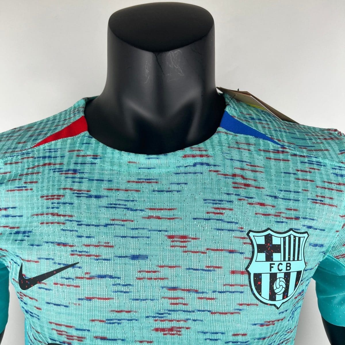 Close-up of a black mannequin torso wearing a turquoise plaid shirt with red and blue accents; a partial FC Barcelona crest is visible on the right.