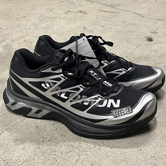 rn-image_picker_lib_temp_de7bfe5c-4229-4f4d-b225-1fe73b4b6a27 | UAE SPORT STORE Pair of black and silver Salomon running shoes with a chunky midsole and attached price/tag on a concrete surface.